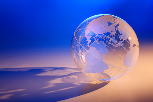 A transparent globe lit with a warm light sitting on a blue background.  A strong directional sidelight casts its shadow across the surface. The globe is rotated to show North America and South America.