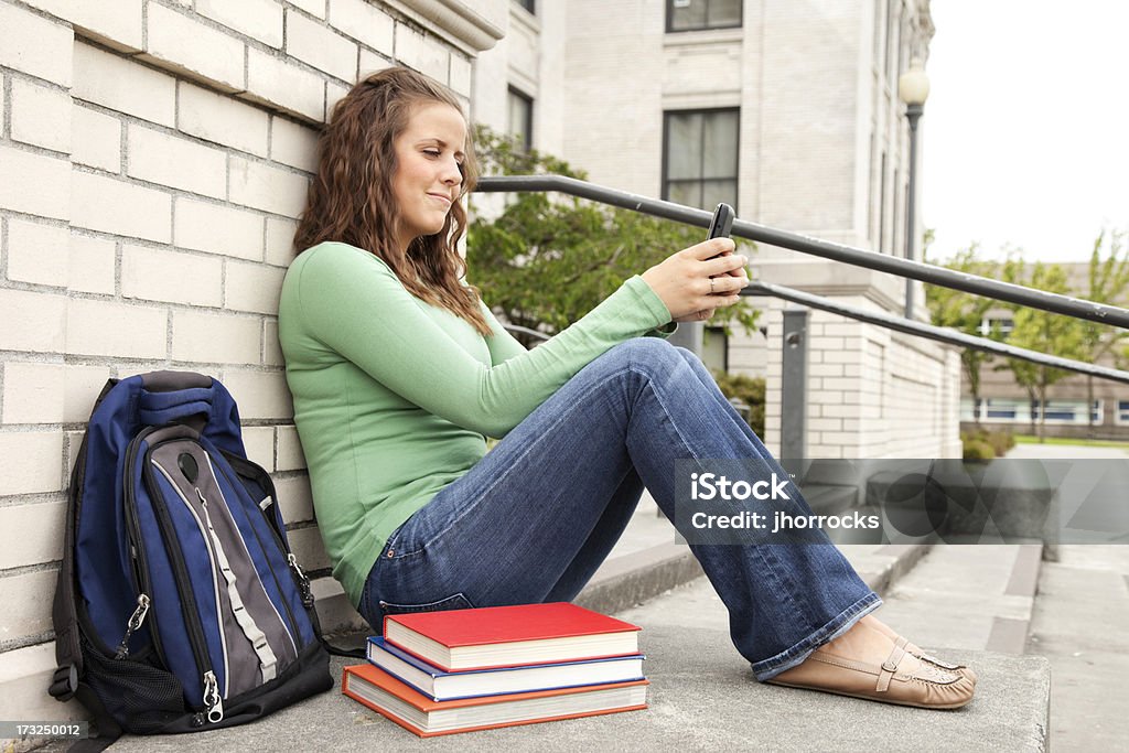 Studente seduto al di fuori del mondo della scuola con libri e telefono cellulare - Foto stock royalty-free di 18-19 anni