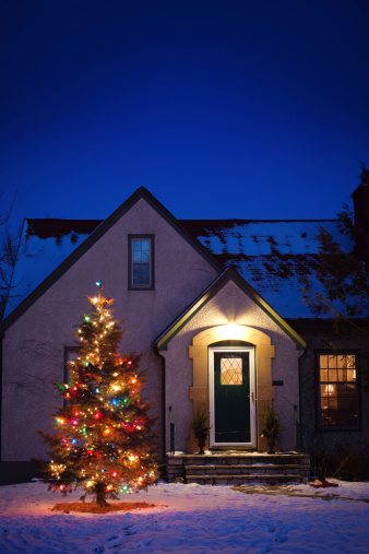 Vertical view of an old-fashioned, small, quaint stucco house with an evergreen tree in its front yard. Photographed in the late dusk light on a winter evening. Roof and yard are dusted in snow. The front door light glows from a warm, welcoming exterior light over the entry, and the decorated tree is covered with multi-color Christmas lights. The dark blue-purple sky allows room for copy space.