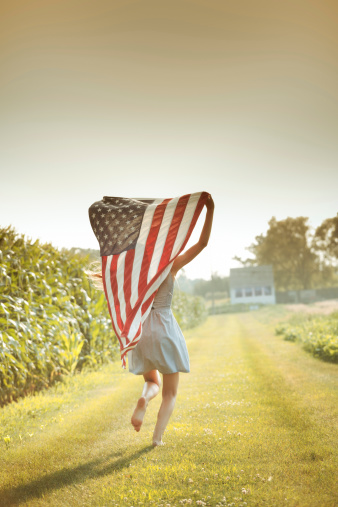 Young farm girl running next to corn crop farm field, holding the USA American flag over her head, flying it behind her. The national banner is displayed for the Fourth of July, Memorial Day, and other political or patriotic holidays and events. Summer light glows on this rural Midwest landscape.