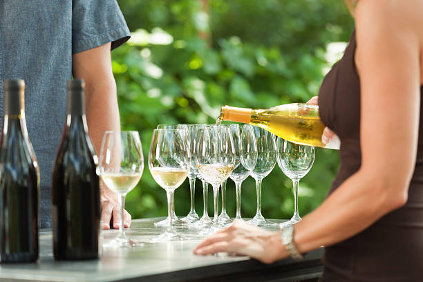 Bartender Pouring White Wine from Bottle for Outdoor Winery Tasting An unidentifiable woman bartender pouring white wine from a wine bottle into wine glasses for outdoor wine tasting at a Napa Valley, California, USA winery. An unidentifiable customer, a wine country vacationing tourist, is served the alcoholic beverage. The counter displays red wine bottles. Vineyard grapevines are in the background. napa california stock pictures, royalty-free photos & images