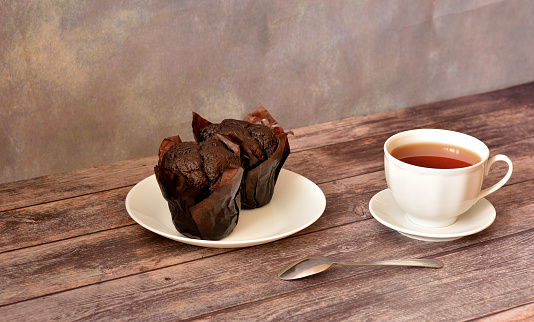 A cup of hot tea on a saucer with a spoon and a plate with two paper-wrapped chocolate muffins on a wooden table. Close-up.