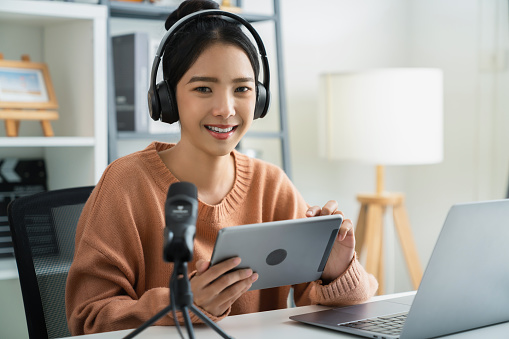 Young Asian woman holding digital tablet and use microphones wear headphones with laptop record podcast interview for radio. Content creator concept.