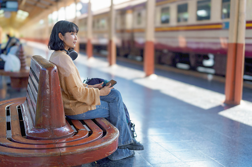 An Asian female tourist, sitting, on a bench, at a train station, wearing headphones, listening to music, from a tablet, while waiting for her train to arrive.