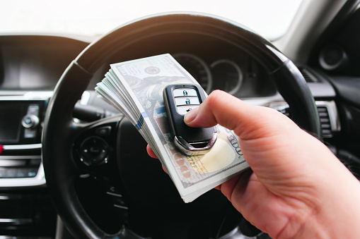 Driver hand holding a car key or keyless remote and usd dollar banknotes with a steering wheel on background in a car. Buy sell and car loan concept