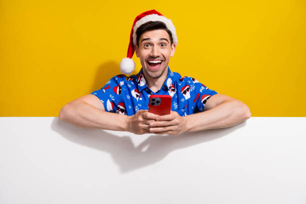Portrait of astonished handsome guy use smart phone empty space x-mas ad placard isolated on yellow color background
