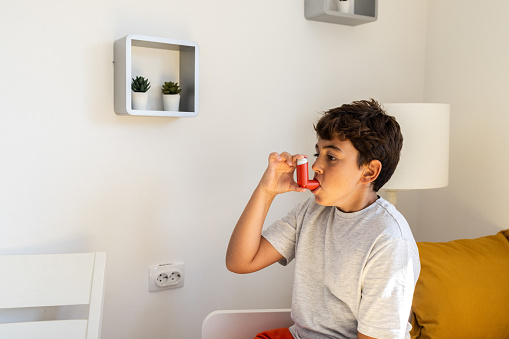 Side-view of an asthmatic Caucasian boy, using an asthma inhaler, while sitting on his bed in a bedroom