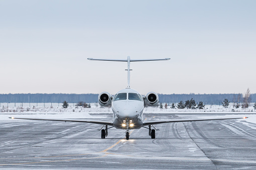 Front view of the modern white business jet taxiing on airport taxiway in winter