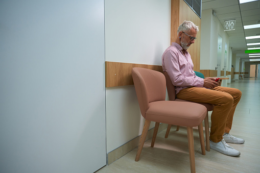 Elderly man sits with a mobile phone in a hospital corridor, the room is bright and clean