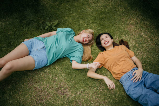 Two female friends, with cheerful smiles, are lying on the park's green lawn.