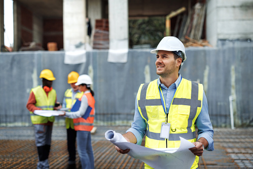 Portrait of male architect with reflective clothes analyzing blueprint while his colleagues are talking in the background