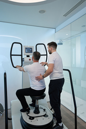 Adult person working out in seated position on multi-axis motorized platform with assistance of physiotherapist
