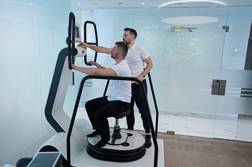 Man seated on stool of multi-axis motorized platform doing balance exercise supervised by experienced physiotherapist on machine touchscreen