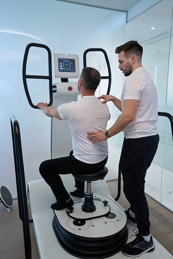 Man exercising in seated position on multi-axis motorized platform with help of physical therapist