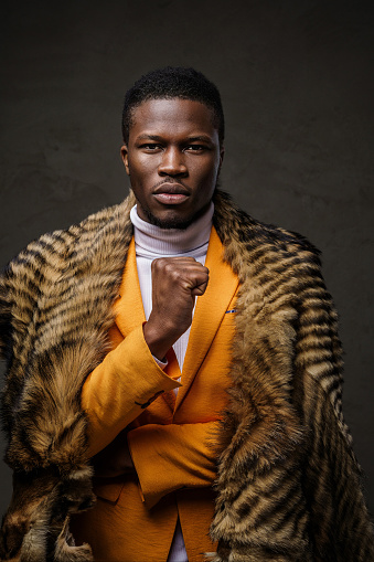Portrait of a stylish dark-skinned man wearing a vibrant yellow jacket with a wild animal pelt draped over his shoulders, confidently posing with a clenched fist against a dark, textured backdrop
