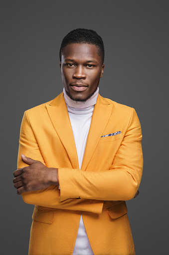 A thoughtful dark-skinned man wearing an elegant yellow jacket and a white turtleneck poses with crossed arms against a gray background
