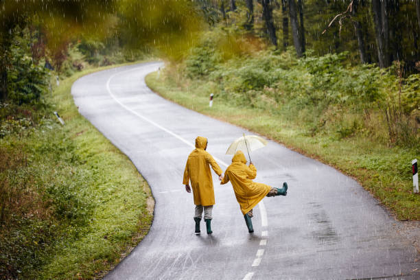 vista posteriore di una coppia spensierata che si diverte sulla strada durante la giornata piovosa. - candid women african descent umbrella foto e immagini stock