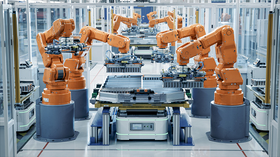 Row of Robot Arms inside Bright Plant Assemble Batteries for Automotive Industry. EV Battery Pack Automated Production Line Equipped with Orange Advanced Robotic Arms. Modern Electric Car Smart Factory