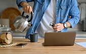 Midsection of businessman pouring hot water in coffee cup while using laptop on kitchen counter