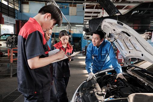 Asian male professional automotive engineer supervisor describes car engine maintenance and repair work with mechanic worker staffs team in fix service garage, specialist occupations in auto industry.