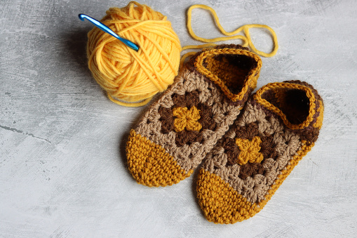 Crocheted slippers made of thick wool and ball of yellow yarn on a gray background with space for text. Granny square pattern close up.