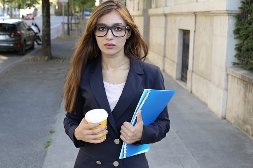 A beautiful South Asian serious woman in her twenties is holding some books and a coffee cup outdoors. She wears eyeglasses with an elegant dark blue blazer.