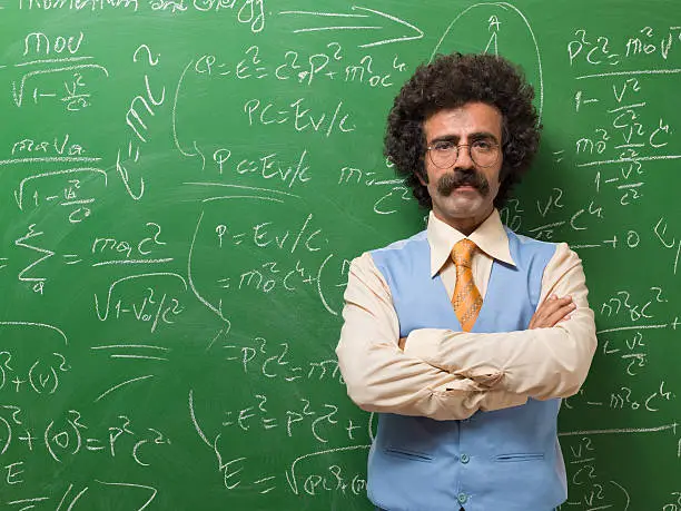 Mature adult teacher wearing a yellow button down shirt,blue waist,pants and necktie standing in front of blackboard for teaching math.He is standing on the right side of frame.The green chalkboard is full of mathematics.He is wearing glasses and has curly hair and mustache.The photo was shot is studio with medium format DSLR camera Hasselblad horizontally.