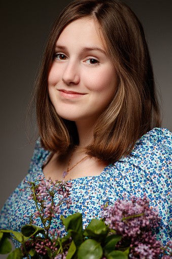 Portrait of beautiful teenage girl with brown hair wearing spring dress, smiling serenely at camera and holding bunch of lilac flowers, studio shot