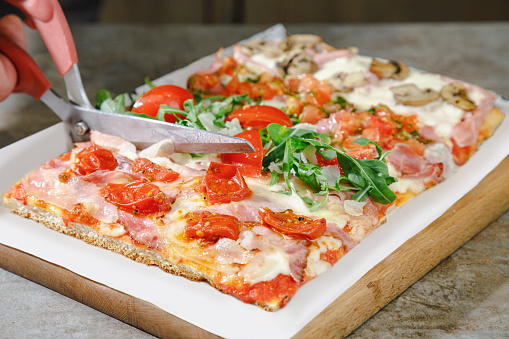 Close-up on pizza with various juicy toppings on one flatbread next to each other in rectangular stripes, cutting with special scissors and serving pizza al taglio