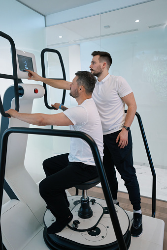 Patient seated on stool of multi-axis motorized platform while physical therapist setting up exercise training program on touchscreen