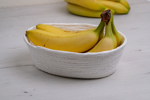 View on Bunch of organic banana fruits is into a white wicker basket on natural wooden background. Close up of Barely ripe yellow bananas in natural wicker container.