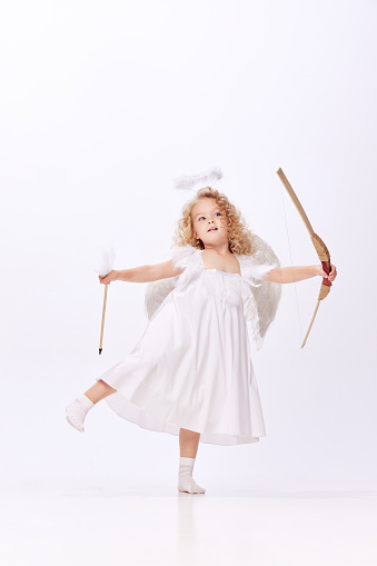 Little beautiful girl, child in image of cupid, angel holding bow isolated over white studio background. Valentines' day. Concept of childhood, imagination, fantasy, fashion and beauty, holidays