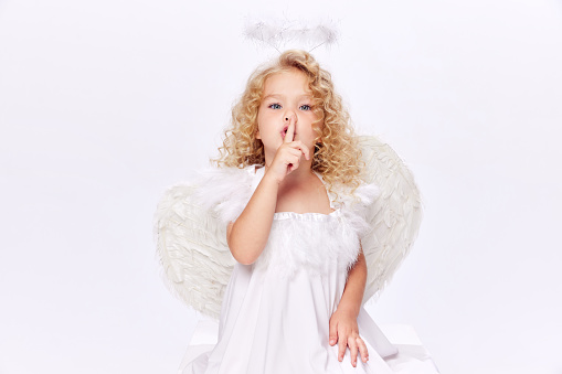 Shh, secret and mystery. Little baby girl in costume of angel holding finger to mouth isolated over white studio background. Concept of childhood, imagination, fantasy, fashion and beauty, holidays