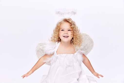 Beautiful, smiling, cute little girl child in white dress in image of angel standing isolated over white studio background. Concept of childhood, imagination, fantasy, fashion and beauty, holidays
