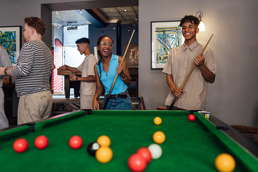 A medium shot of a multiracial group of students out in a pub in Newcastle Upon Tyne. Two of them play pool as their friends stand and talk with drinks.