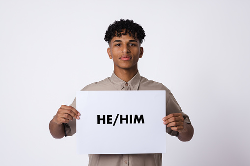 A studio, portrait shot of a male student wearing casual clothing on a white background. He is holding a white sign displaying the words he/him on it.