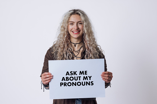 A portrait shot of a non-binary student, with blond curly hair and wearing casual hipster clothing. They are looking and smiling at the camera as they hold a white sign displaying the words he/him on it.