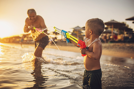 Man and grandson playing with squirt guns in the sea in sunset, they are on vacation.