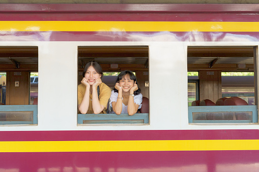 Two Asian female tourists are resting their chins on their hands and smiling broadly out the train window.