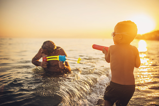 Man and grandson playing with squirt guns the sea in sunset, they are on vacation.