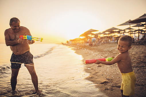 Man and grandson playing with squirt guns by the sea in sunset, they are on vacation.