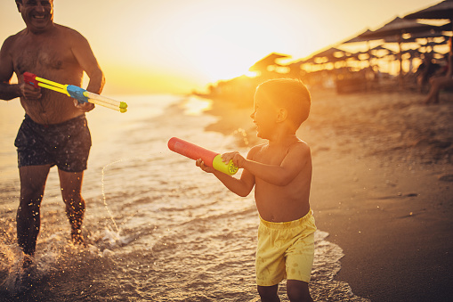 Man and grandson playing with squirt guns by the sea in sunset, they are on vacation.