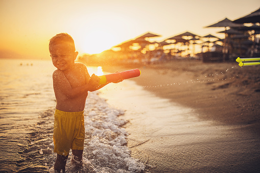 Little boy playing with squirt gun standing on the beach in the sea.