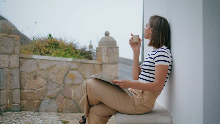 Woman reading morning newspaper on terrace. Relaxed girl drink coffee outdoors