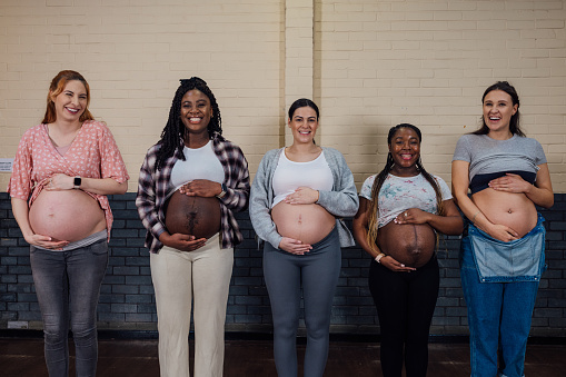 A medium close up front view of a group of women who are enjoying their pregnancies. They are all in their third trimester and are all looking into the camera holding their bumps as they stand in a community centre in the North East of England