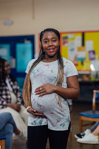 A medium close up selective focus portrait of a 27 weeks pregnant woman with braided hair in the 3rd trimester of her pregnancy she is looking into the camera as she attends an antenatal class that also does fitness sessions in a community centre in the North East of England.