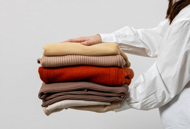 Woman hands holding stack warm winter sweaters on grey background. A stack of sweaters, jumpers in female hands. Winter and autumn warm cozy sweaters for charity.