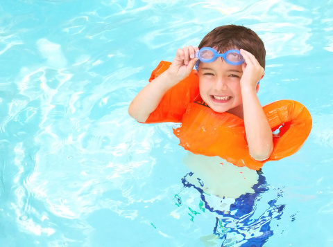 A young boy swimming in a life vest and goggles stops to smile.