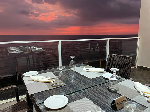 Close-up image of glass topped square tables with rattan wickerwork chairs on hotel balcony with glass safety barrier, tables set ready for meal, dramatic atmospheric sunset background, focus on foreground