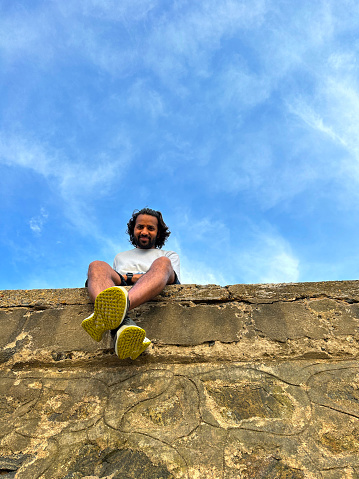 Stock photo showing close-up view of an Indian man sitting on top of manmade, sea defence concrete, seawall, which has been constructed to prevent flooding of a promenade during high tide.